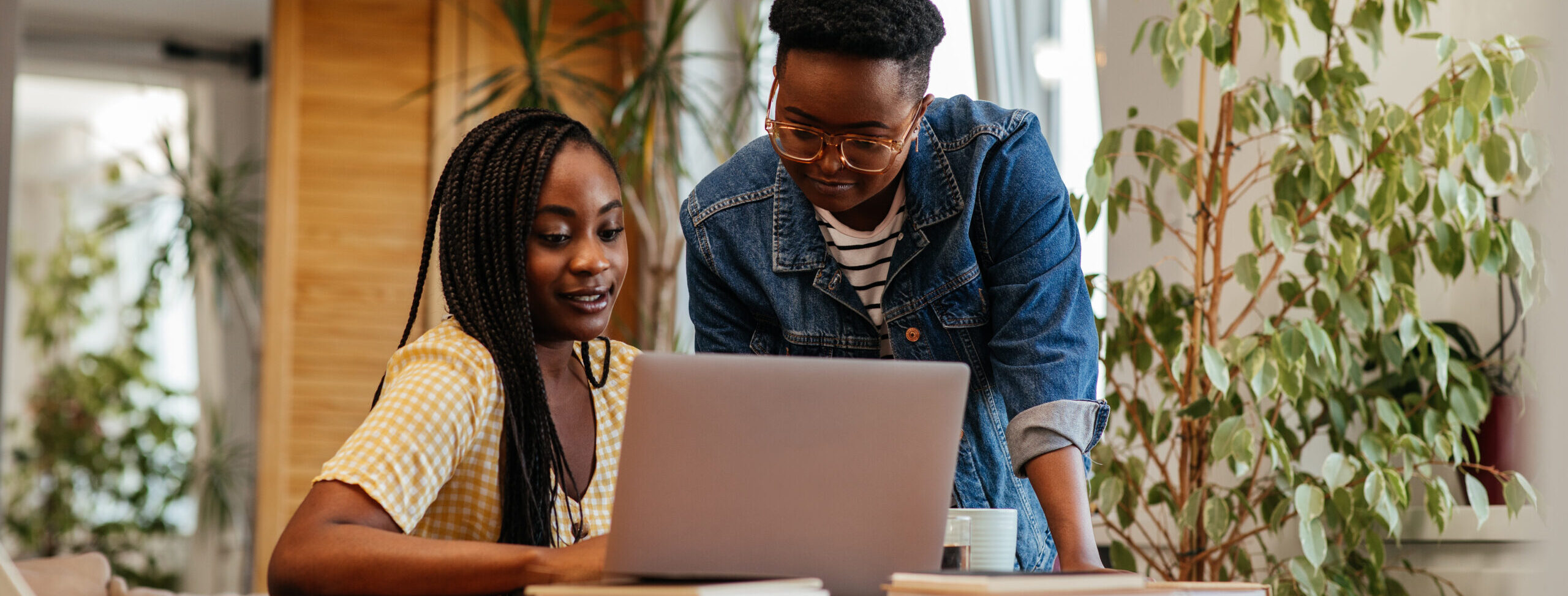 Two black woman looking at a laptop together. They are at a wooden desk with foliage around them.