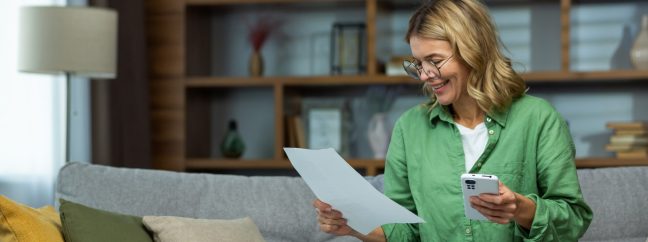 An elderly smilling oman sits at home on the sofa, holds the phone in her hands, works with documents, financial accounts, rent, loans, agreements.