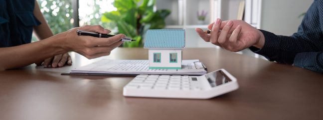 Hands engaged in a discussion over a real estate contract and a miniature house model, representing the negotiation and buying process.