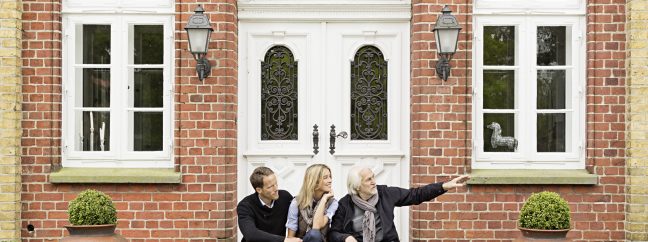 A young adult couple sitting on steps of a brick house with a white door and white windows with an older man.