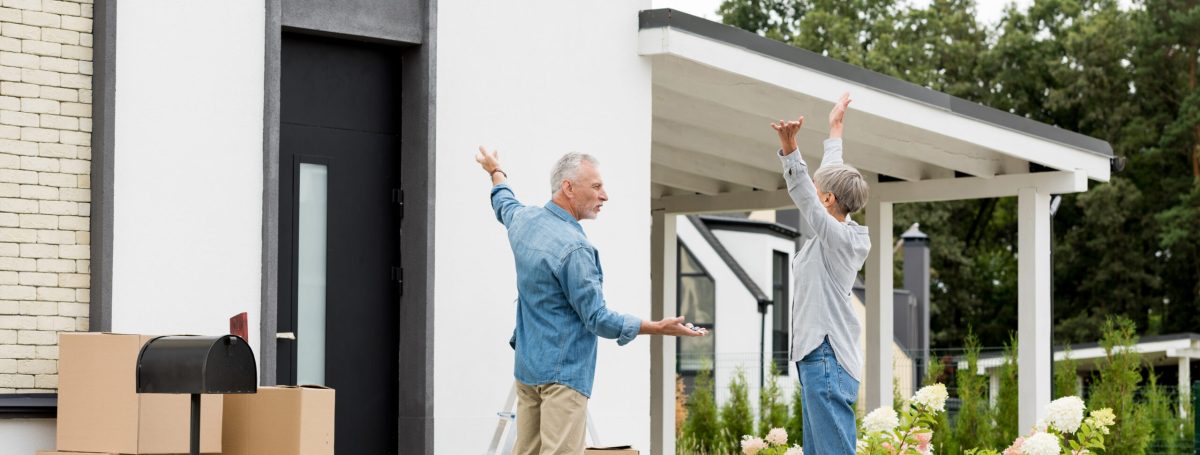 Mature man pointing in with hand and holding keys of new house to woman with outstretched hands.