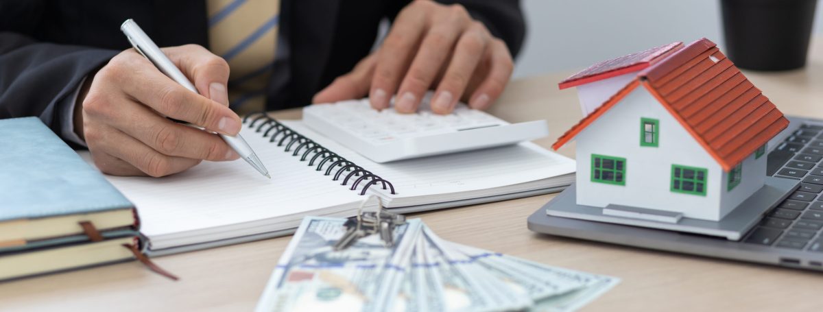 Real estate agent writing in notebook with a money spread and home diorama in front of them.