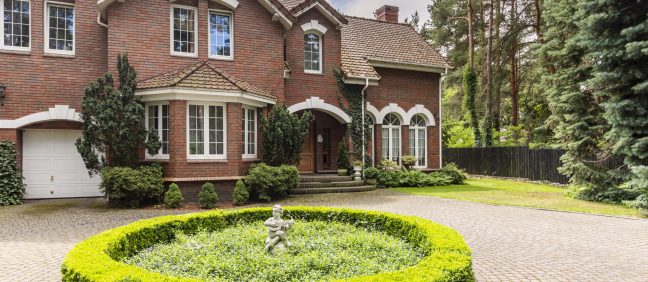 Round hedge and a small statue in the middle of a cobblestone driveway to an elegant, english style house with white windows and brick exterior