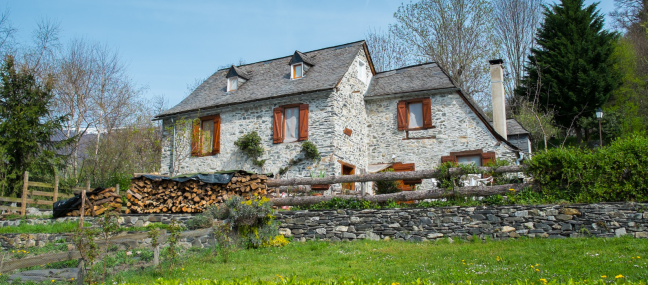 A dutch-style house with a white, exposed brick exterior. There is a pile of firewood in front with a stone fence.