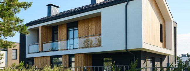Two storey cottage with balcony and group of windows surrounded by green vegetation growing along fencing