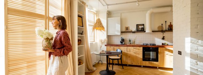 Woman walks with a vase of flowers, decoration and having some house chores at home. Wide interior view on sunny studio apartment in beige tones