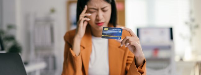 A young woman wearing a white shirt and yellow jacket rubbing her forehead looking unhappy while holding a credit card.